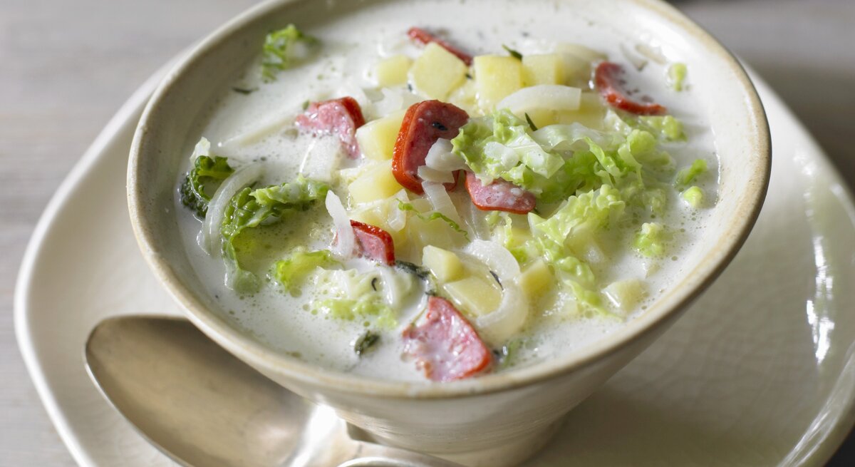 In einer beigefarbenen Suppenschale steht eine cremige Wirsingsuppe mit Wurst. In der hellen Brühe schwimmen Stücke von grünem Wirsing, gelben Kartoffelwürfeln und roten Wurstscheiben. Die Suppe steht auf einem Teller, daneben liegt ein Löffel. Im Hintergrund ist ein unscharfer Holztisch zu erkennen, der der Szene eine warme, gemütliche Atmosphäre verleiht.