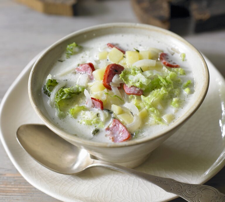 In einer beigefarbenen Suppenschale steht eine cremige Wirsingsuppe mit Wurst. In der hellen Brühe schwimmen Stücke von grünem Wirsing, gelben Kartoffelwürfeln und roten Wurstscheiben. Die Suppe steht auf einem Teller, daneben liegt ein Löffel. Im Hintergrund ist ein unscharfer Holztisch zu erkennen, der der Szene eine warme, gemütliche Atmosphäre verleiht. | © Stockfood/Cimbal, Walter