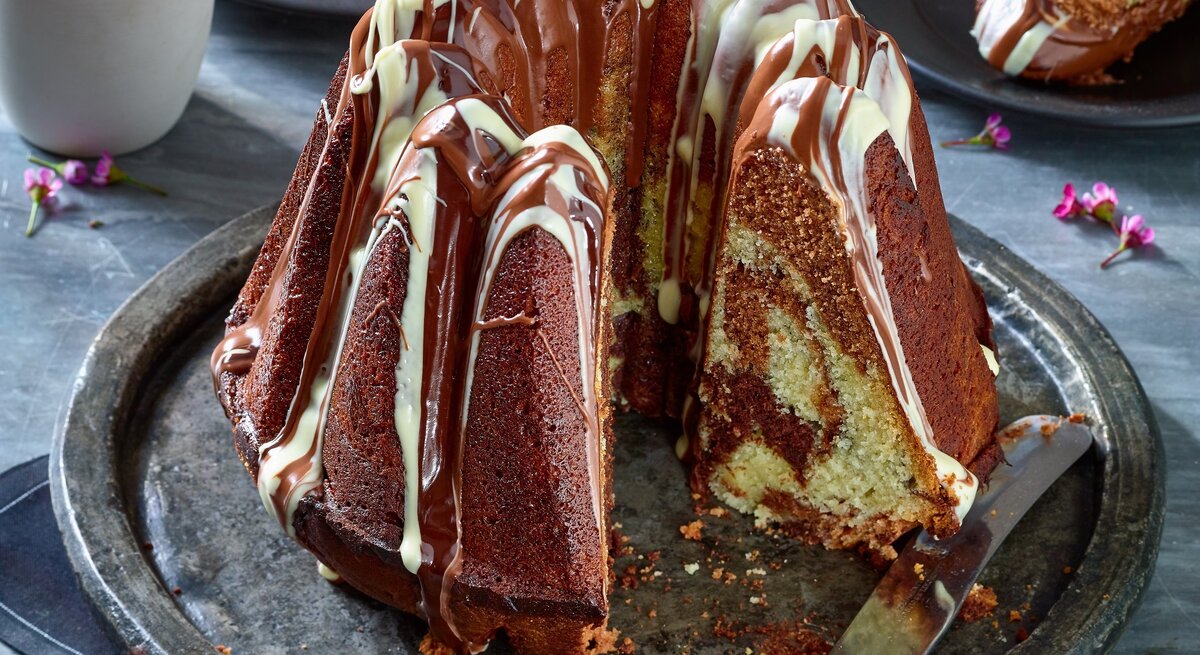 Ein saftiger Marmorkuchen mit Schokoladenglasur und heller Verzierung steht auf einem silbernen Teller, ein Stück wurde herausgeschnitten.