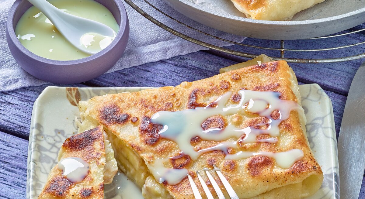 Der Bananen-Pfannenstrudel auf einem eckigen hellen Teller mit heller Soße. Alles auf einem blau-violetten Untergrund.