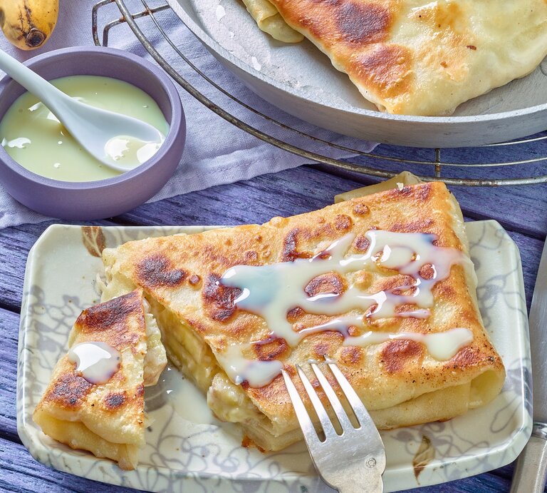 Der Bananen-Pfannenstrudel auf einem eckigen hellen Teller mit heller Soße. Alles auf einem blau-violetten Untergrund. | © EAT CLUB/ Matthias Liebich
