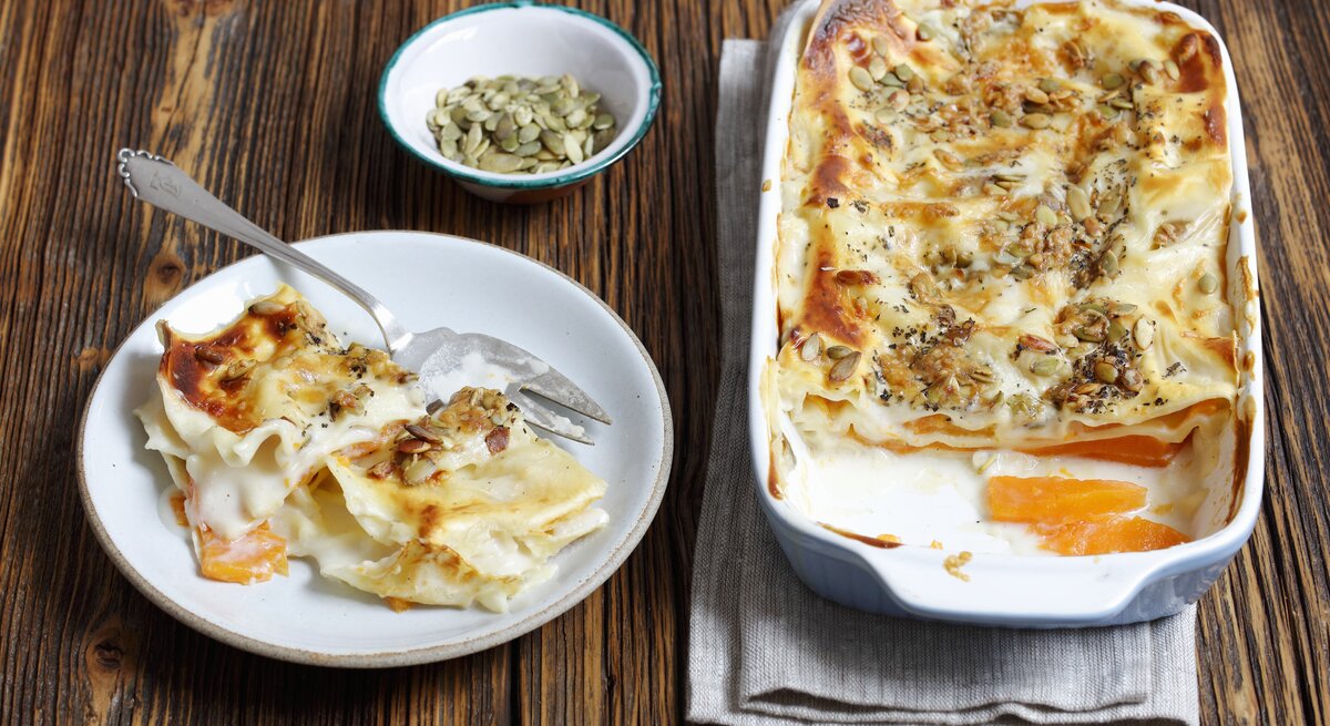 Die vegetarische Kürbis-Lasagne mit Kürbiskernen auf einem dunklen Holztisch von schräg oben fotografiert.