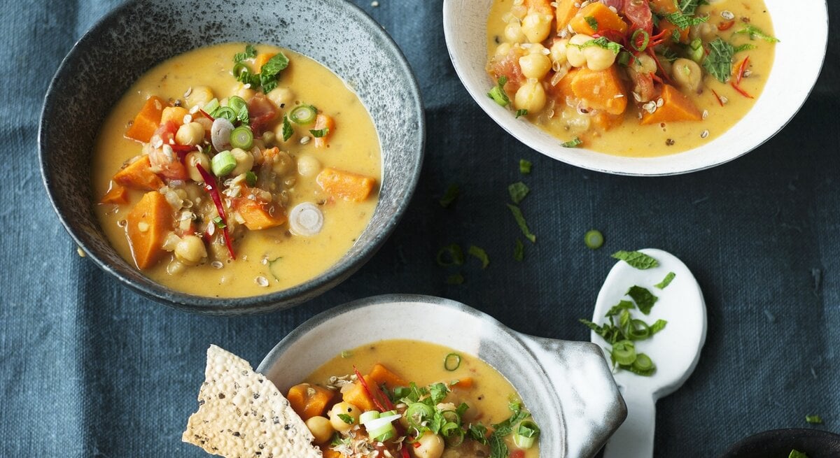 Das Bild zeigt drei Schüsseln, gefüllt mit einer cremigen Suppe, die Stücke von Gemüse und Kichererbsen enthält. Neben den Schüsseln liegen dünne, knusprige Brotscheiben und ein kleiner Teller mit einem Löffel. Im Vordergrund steht eine kleine Schale mit frisch geschnittenen Kräutern. Die Szene vermittelt den Eindruck einer liebevollen und vielseitigen Mahlzeit.