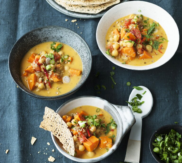 Das Bild zeigt drei Schüsseln, gefüllt mit einer cremigen Suppe, die Stücke von Gemüse und Kichererbsen enthält. Neben den Schüsseln liegen dünne, knusprige Brotscheiben und ein kleiner Teller mit einem Löffel. Im Vordergrund steht eine kleine Schale mit frisch geschnittenen Kräutern. Die Szene vermittelt den Eindruck einer liebevollen und vielseitigen Mahlzeit. | © seasons.agency / Grfe & Unzer Verlag / Schardt, Wolfgang
