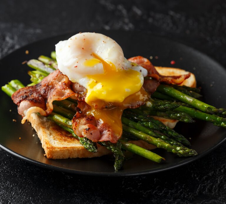 Mit Spargel, Speck und Ei belegtes Toastbrot auf schwarzem Teller.  | © Getty Images/ DronG