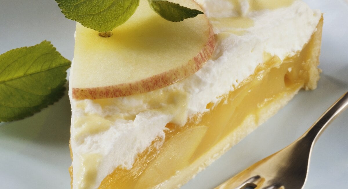 Nahaufnahme eines Stücks Eierlikör-Apfel-Torte mit Vanillecreme, frischen Apfelscheiben und Minzblättern auf einem weißen Teller, daneben eine Kuchengabel