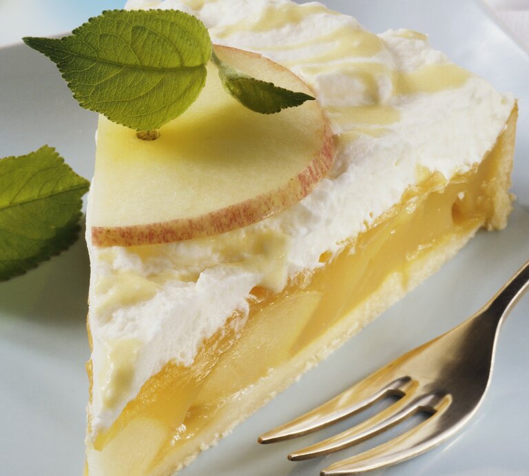 Nahaufnahme eines Stücks Eierlikör-Apfel-Torte mit Vanillecreme, frischen Apfelscheiben und Minzblättern auf einem weißen Teller, daneben eine Kuchengabel | © StockFood