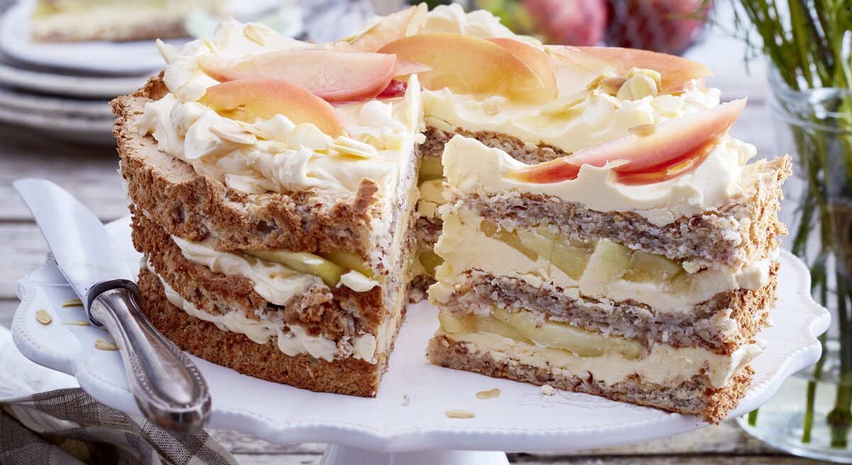 Schwedische Apfel-Mandeltorte angeschnitten auf einem weißen Tortenteller, darauf liegt ein Messer und links davon liegt ein Geschirrtuch. Der Hintergrund ist hölzern und hinten auf dem Foto eine Schüssel mit Äpfeln und kleine Teller gestapelt - darauf ein Stück Torte.
