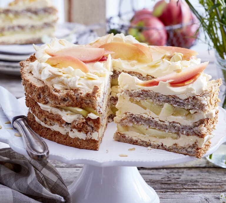 Schwedische Apfel-Mandeltorte angeschnitten auf einem weißen Tortenteller, darauf liegt ein Messer und links davon liegt ein Geschirrtuch. Der Hintergrund ist hölzern und hinten auf dem Foto eine Schüssel mit Äpfeln und kleine Teller gestapelt - darauf ein Stück Torte. | © EAT CLUB / Isabell Triemer