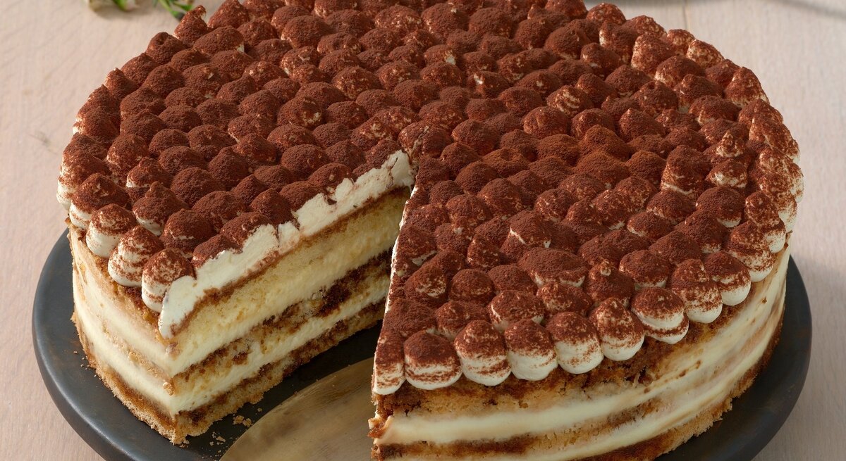 Die angeschnittene Kaffee-Schoko-Torte auf einem dunklen Tortenständer und beigem Holztisch. Alles von schräg oben fotografiert.