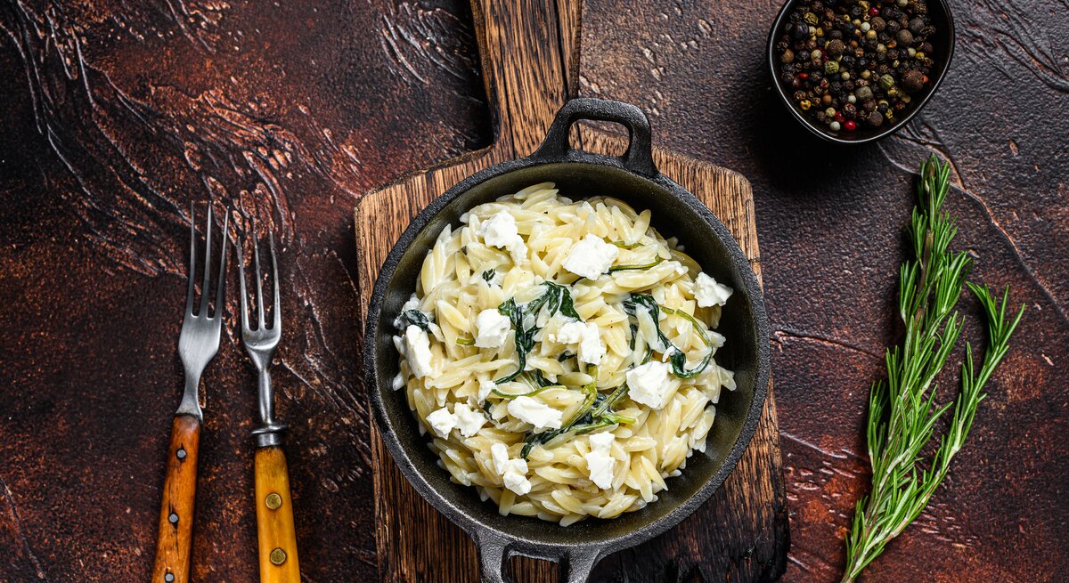 Gusseiserne Pfanne gefüllt mit Nudelreis, Spinat und Käse in rustikalem Ambiente mit mediterraner Dekoration.