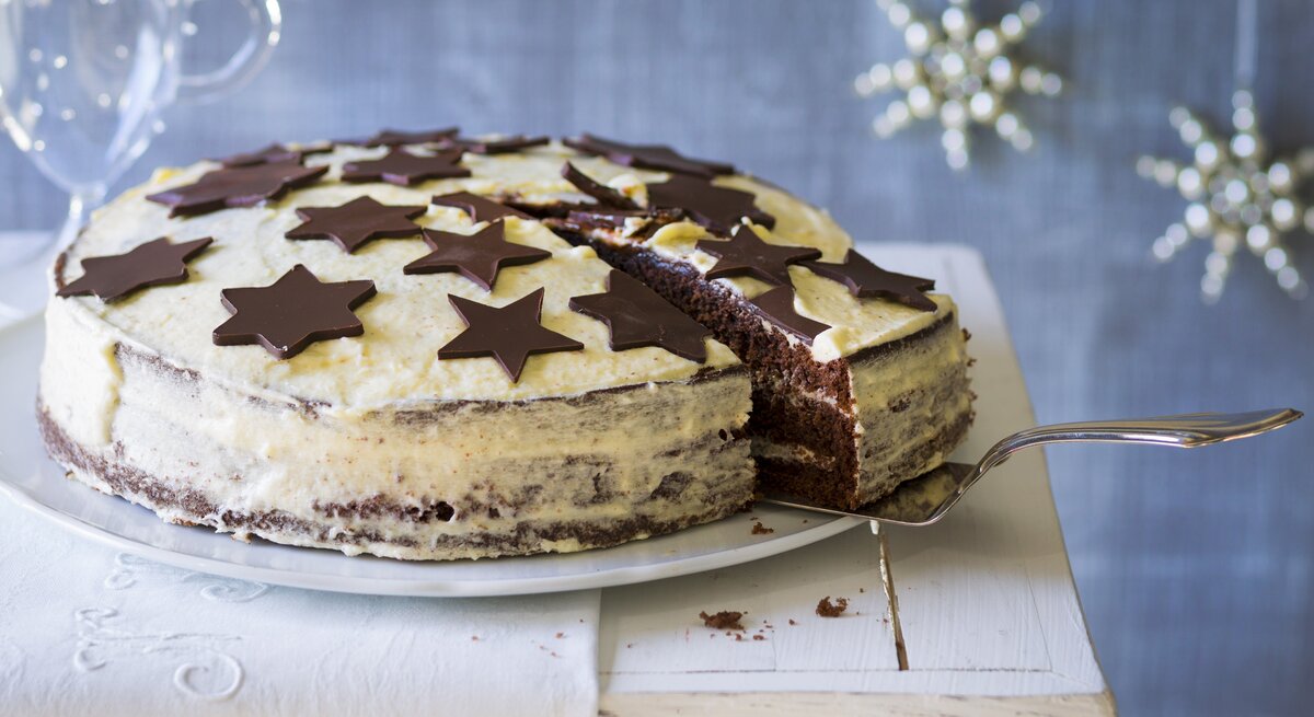 Weißer Kuchen mit schokoladigen Sternen vor weihnachtlich geschmücktem, blauen Hintergrund. 