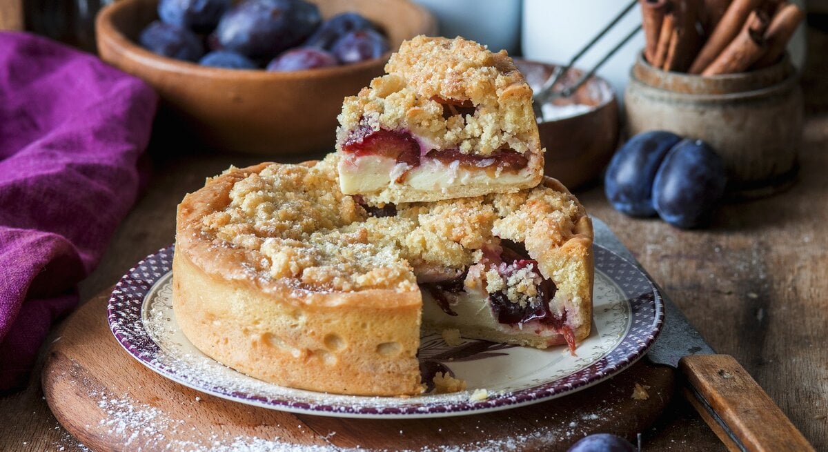 Der Streusel-Zwetschgen-Quarkkuchen auf einem rustikalen Holztisch mit Zwetschgen im Hintergrund fotografiert.