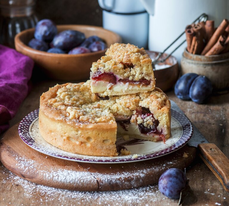 Der Streusel-Zwetschgen-Quarkkuchen auf einem rustikalen Holztisch mit Zwetschgen im Hintergrund fotografiert. | © StockFood/Meliukh, Irina