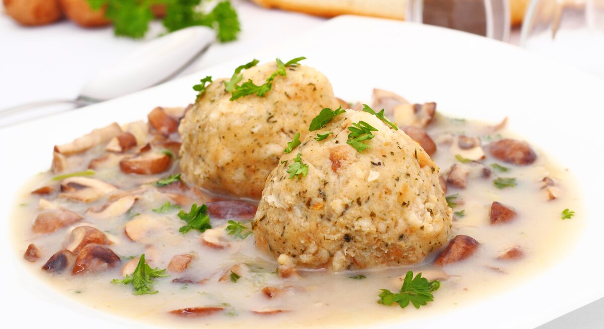 Auf einem weißen Teller liegen zwei Semmelknödel, garniert mit frischer Petersilie, umgeben von einem cremigen Ragout mit kleinen Pilzstücken. Im Hintergrund sind Brot, Kräuter und Geschirr zu erkennen.
