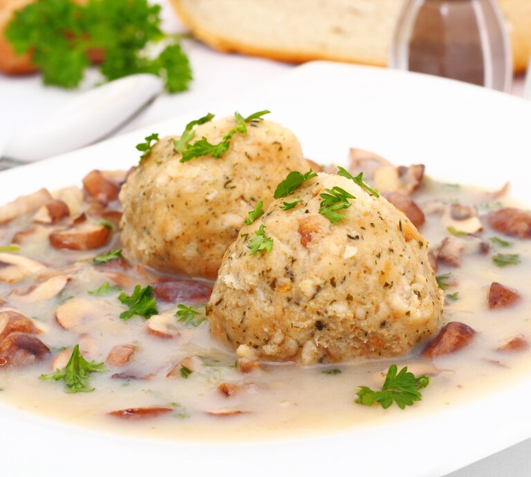 Auf einem weißen Teller liegen zwei Semmelknödel, garniert mit frischer Petersilie, umgeben von einem cremigen Ragout mit kleinen Pilzstücken. Im Hintergrund sind Brot, Kräuter und Geschirr zu erkennen. | © iStock/vertmedia