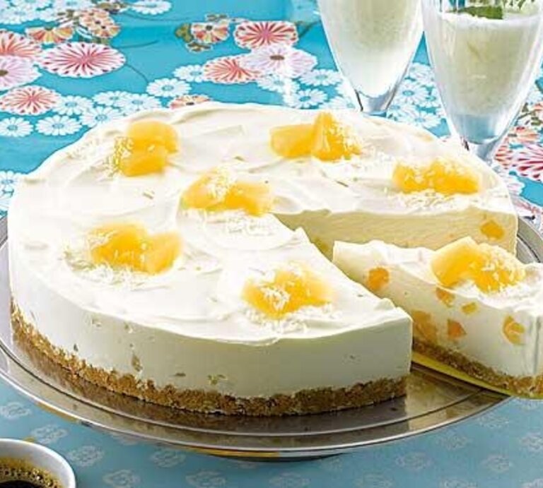 Pina-Colada-Torte mit Ananas-Stücken / Angeschnitten / 1 Stück Torte auf Tortenheber /  Zwei gefüllte Cocktailgläser / Untergrund Tisch / Draufsicht schäg | © Philadelphia