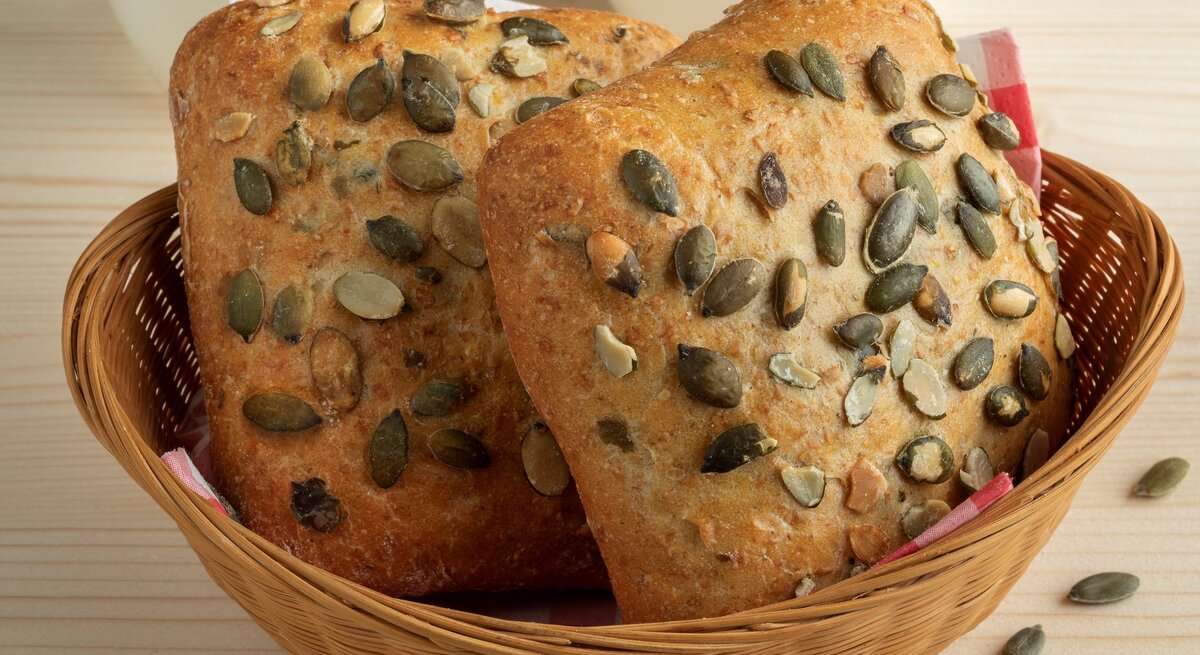 Selbst gemachte Kürbiskernbrötchen in Brotkorb auf Holztisch. Im Bild zwei weiße Tassen und einige Kürbiskerne. Frontalansicht.