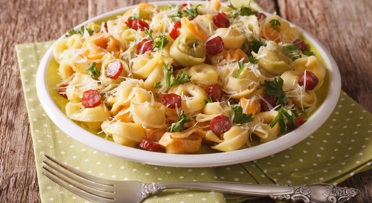 Teller mit Tortellini-Salat mit Salami, Kräutern und Parmesan auf hölzernem Untergrund. 