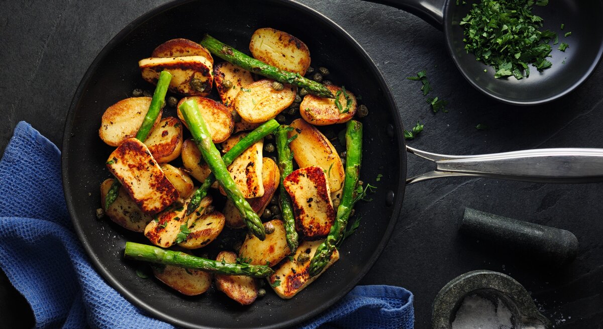 Spargel-Halloumi-Pfanne mit Kartoffeln in gusseiserner Pfanne auf dunklem Untergrund. 