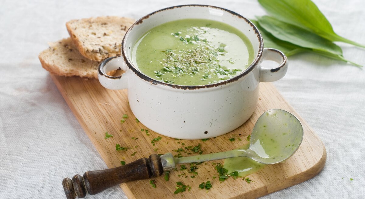 Ein Topf mit cremiger Bärlauchsuppe, bestreut mit Kräutern, neben einem Stück Brot auf einem Holzbrett, mit einem alten Löffel und frischen Bärlauchblättern daneben.
