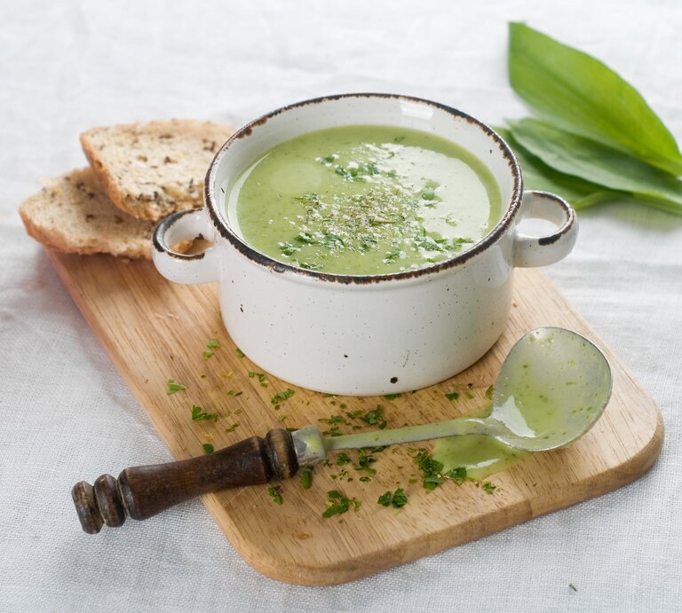 Ein Topf mit cremiger Bärlauchsuppe, bestreut mit Kräutern, neben einem Stück Brot auf einem Holzbrett, mit einem alten Löffel und frischen Bärlauchblättern daneben. | © iStock/ Wiktory