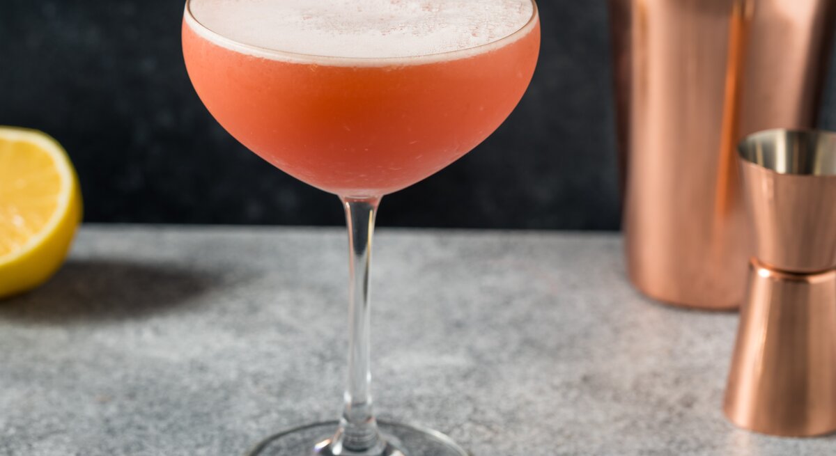 Negroni Sour in einer Coupette mit Shaker, Barmaß und einer Zitrone vor grauem Hintergrund. Frontalansicht.