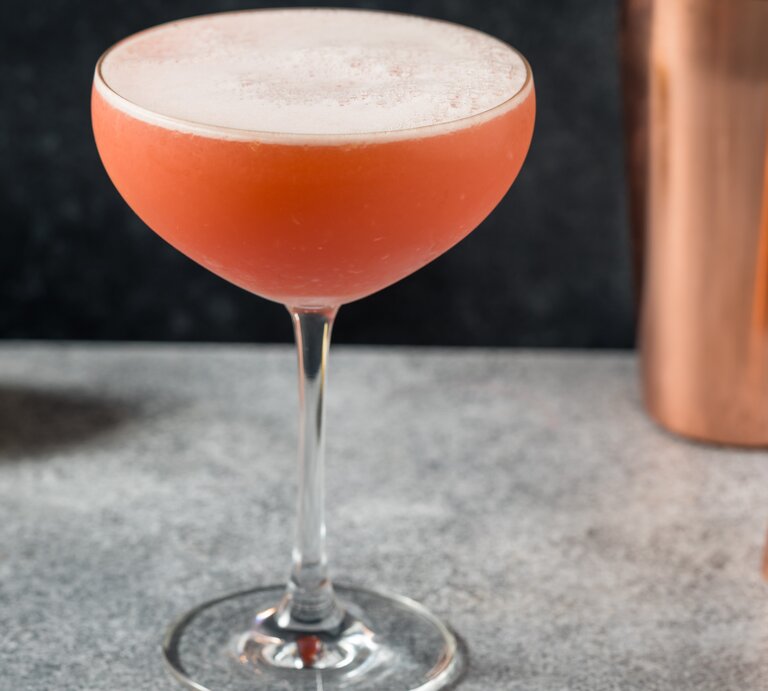 Negroni Sour in einer Coupette mit Shaker, Barmaß und einer Zitrone vor grauem Hintergrund. Frontalansicht. | © Shutterstock / Brent Hofacker