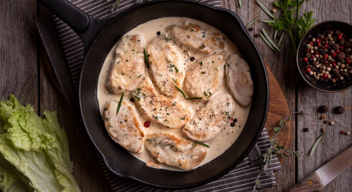 Hähnchen in mexikanischer Sahnesoße in Gusspfanne auf Tischtuch und Holztisch. Im Bild eine Schale mit buntem Pfeffer, Kräutern und Salat. Draufsicht.