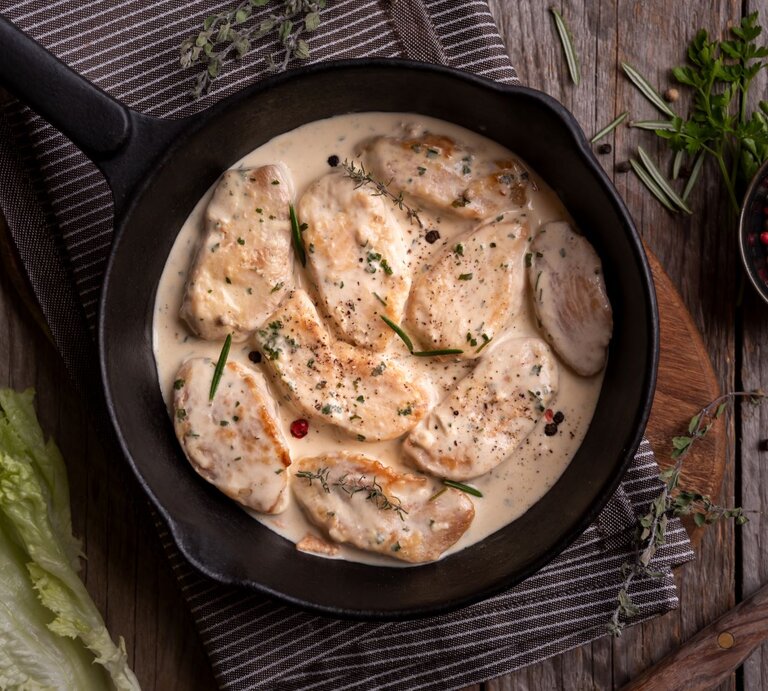 Hähnchen in mexikanischer Sahnesoße in Gusspfanne auf Tischtuch und Holztisch. Im Bild eine Schale mit buntem Pfeffer, Kräutern und Salat. Draufsicht. | © shutterstock / Maria_Usp