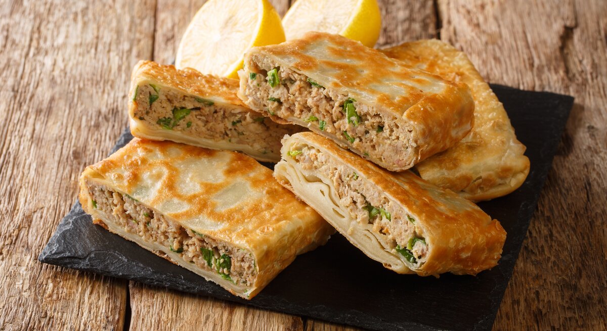 Eckig gefaltete Pfannkuchentaschen mit saftiger Hackfleisch-Füllung auf Schieferplatte. Diese steht auf einem rustikalen Holztisch. 