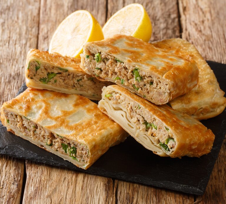 Eckig gefaltete Pfannkuchentaschen mit saftiger Hackfleisch-Füllung auf Schieferplatte. Diese steht auf einem rustikalen Holztisch.  | © GettyImages/ALLEKO