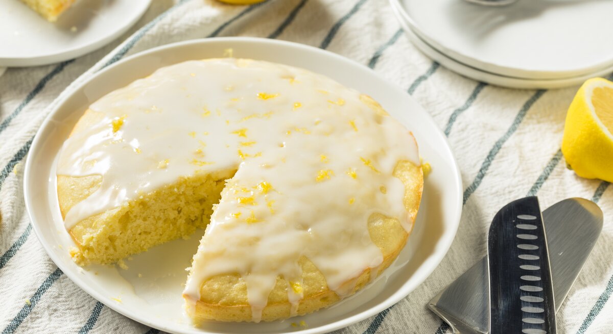 Runder, angeschnittener Zitronenkuchen mit heller Glasur auf einem weißen Teller. 