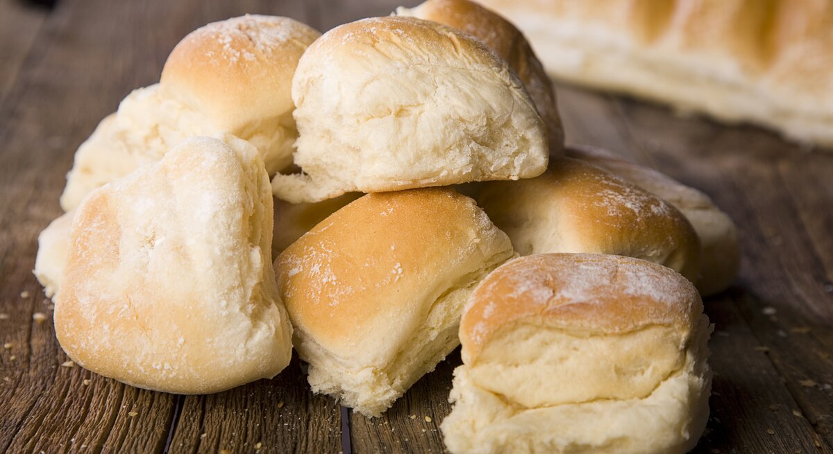 Mehrere goldbraune Quarkbrötchen sind auf einem Holztisch gestapelt, mit einem weiteren Brotlaib im Hintergrund.