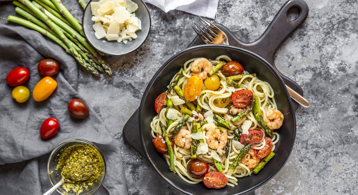 Gusseiserne Pfanne gefüllt mit Spargel-Garnelen-Pasta mit frischen Tomaten und Parmesan auf Marmor-Untergrund. 