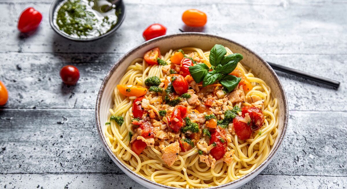 Spaghetti mit Lachs-Sahne-Soße und Bärlauch-Pesto auf Pasta-Teller auf grauem Untergrunmd. Im Bild Tomaten und eine Schale mit Pesto. Draufsicht.