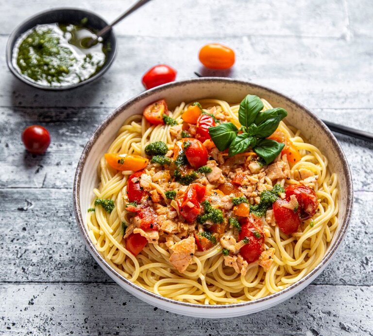 Spaghetti mit Lachs-Sahne-Soße und Bärlauch-Pesto auf Pasta-Teller auf grauem Untergrunmd. Im Bild Tomaten und eine Schale mit Pesto. Draufsicht. | © Getty Images / Westend61