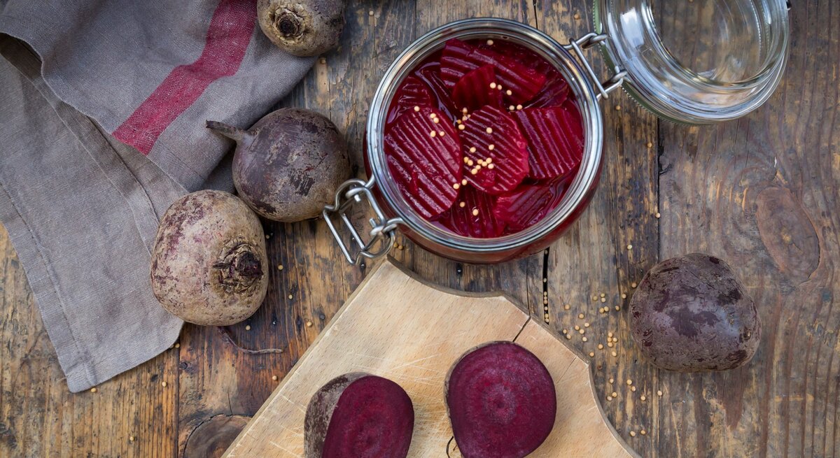 Rote Bete in einem Glas mit geöffnetem Decken. Im Bild eine aufgeschnittene Rote Bete auf Holzbrett. Weitere im Bild verteilt. Arrangiert auf Holztisch. Vogelperspektive.