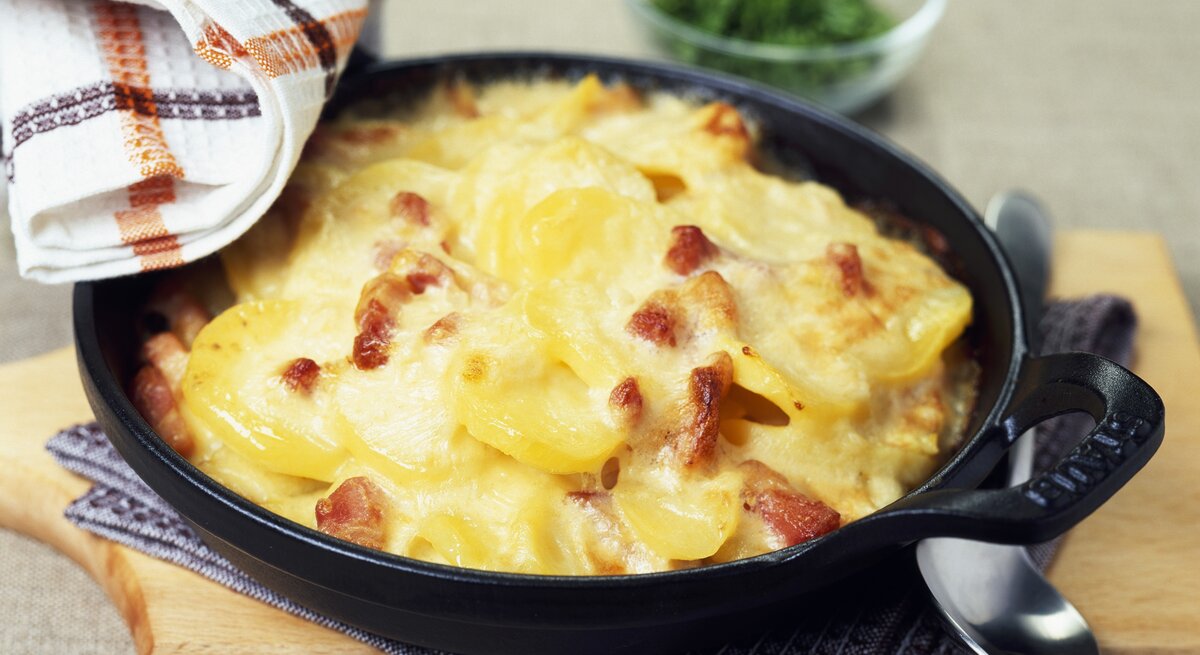 Kartoffelauflauf mit Schinken und Käse in gusseisernen Form auf Holzbrett.