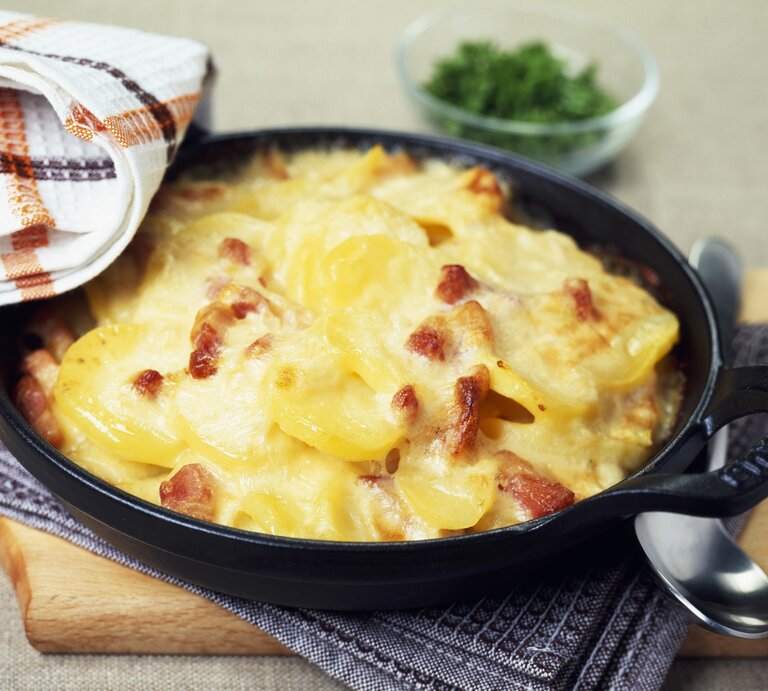 Kartoffelauflauf mit Schinken und Käse in gusseisernen Form auf Holzbrett. | © GettyImages/Norris, Bob