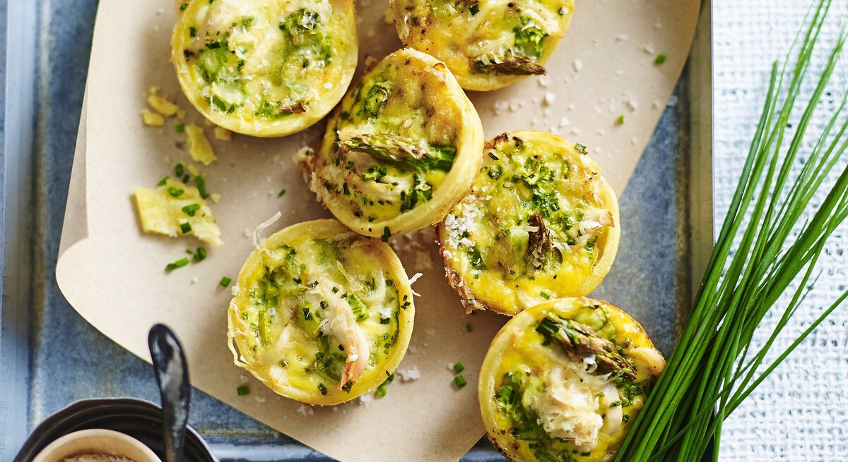 Die Miniquiches mit Hähnchen und Spargel auf einem Blech und weißen Tisch. Alles direkt von oben fotografiert.