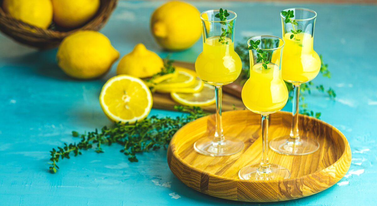Mehrere Gläschen gefüllt mit Limoncello auf einem Holzbrett. Dahinter liegen mehrere Zitronen auf blauem Untergrund. 