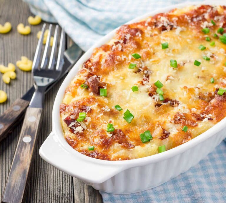 Nudelauflauf Flammkuchen Art in weißer Auflaufform auf blau-weißem Tuch auf Holz. Daneben Besteck und rohe Nudeln. Draufsicht. | © Shutterstock / iuliia_nrez