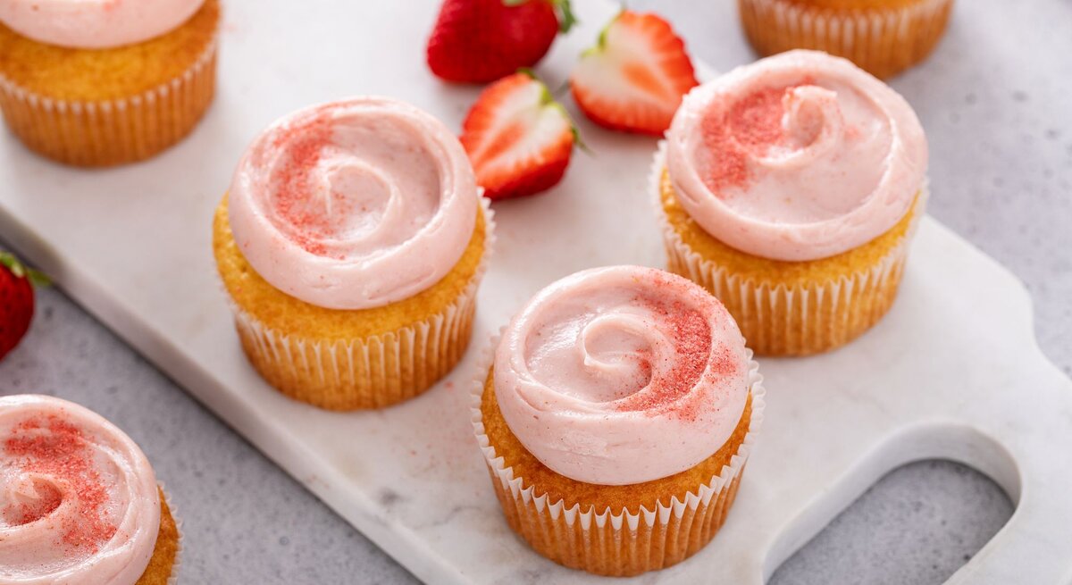 Erdbeer-Zitronen-Cupcakes mit zweifarbig rosa-weiß geschwungenem Frischkäse-Frosting, dekoriert mit frischen Erdbeerscheiben und Zitronenmelisse vor hellem Hintergrund.
