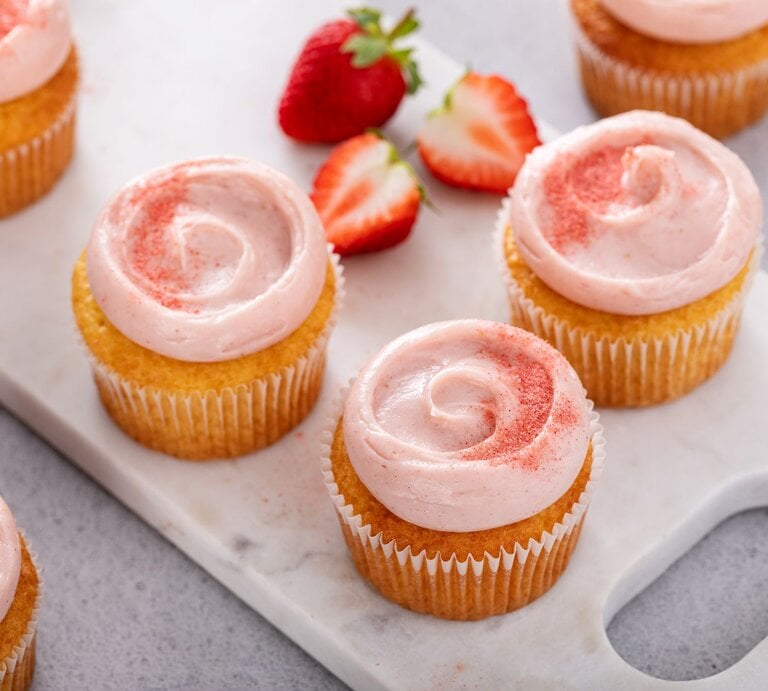 Erdbeer-Zitronen-Cupcakes mit zweifarbig rosa-weiß geschwungenem Frischkäse-Frosting, dekoriert mit frischen Erdbeerscheiben und Zitronenmelisse vor hellem Hintergrund. | © Shutterstock/Elena Veselova