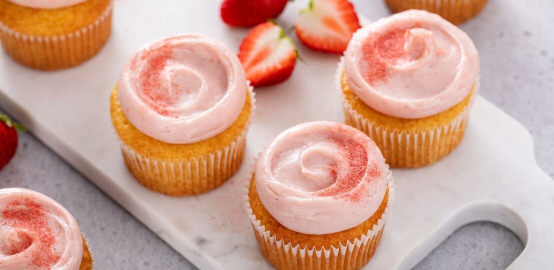 Erdbeer-Zitronen-Cupcakes mit zweifarbig rosa-weiß geschwungenem Frischkäse-Frosting, dekoriert mit frischen Erdbeerscheiben und Zitronenmelisse vor hellem Hintergrund. | © Shutterstock/Elena Veselova