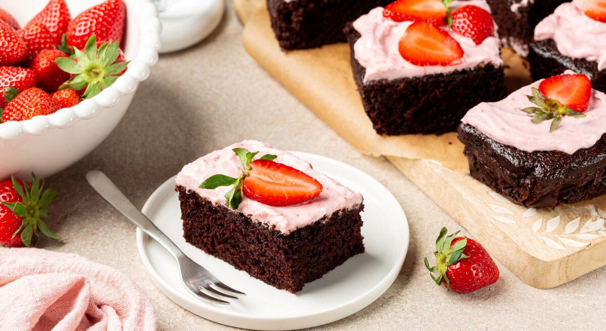 Ein quadratisch geschnittenes Stück sahnige Erdbeer-Brownies auf einem Kuchenteller. Unten ein dunkler, kompakter Schokoboden, darauf eine rosa Erdbeer-Sahneschicht, verziert mit einer frischen Erdbeere. | © Shutterstrock/OlgaBombologna