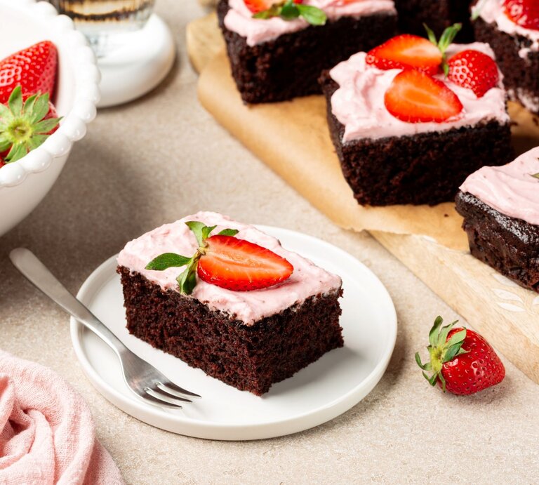 Ein quadratisch geschnittenes Stück sahnige Erdbeer-Brownies auf einem Kuchenteller. Unten ein dunkler, kompakter Schokoboden, darauf eine rosa Erdbeer-Sahneschicht, verziert mit einer frischen Erdbeere. | © Shutterstrock/OlgaBombologna