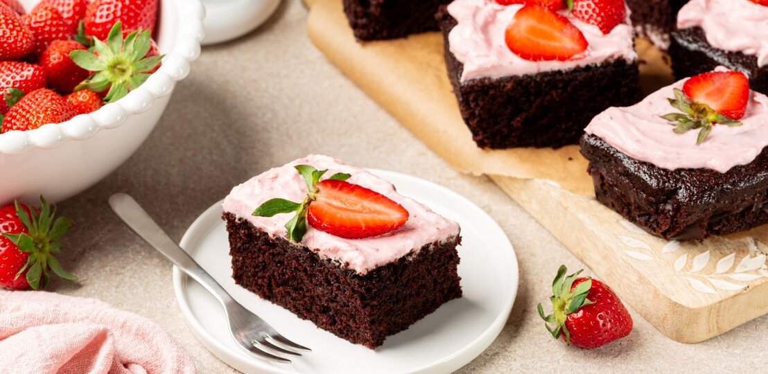 Ein quadratisch geschnittenes Stück sahnige Erdbeer-Brownies auf einem Kuchenteller. Unten ein dunkler, kompakter Schokoboden, darauf eine rosa Erdbeer-Sahneschicht, verziert mit einer frischen Erdbeere. | © Shutterstrock/OlgaBombologna