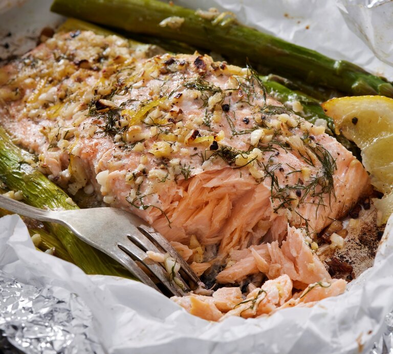 Ofenlachs mit grünem Spargel, Zitrone, Knoblauch und Dill in Backpapier und Alufolie mit einer Gabel. Draufsicht. | © Getty Images / LauriPatterson